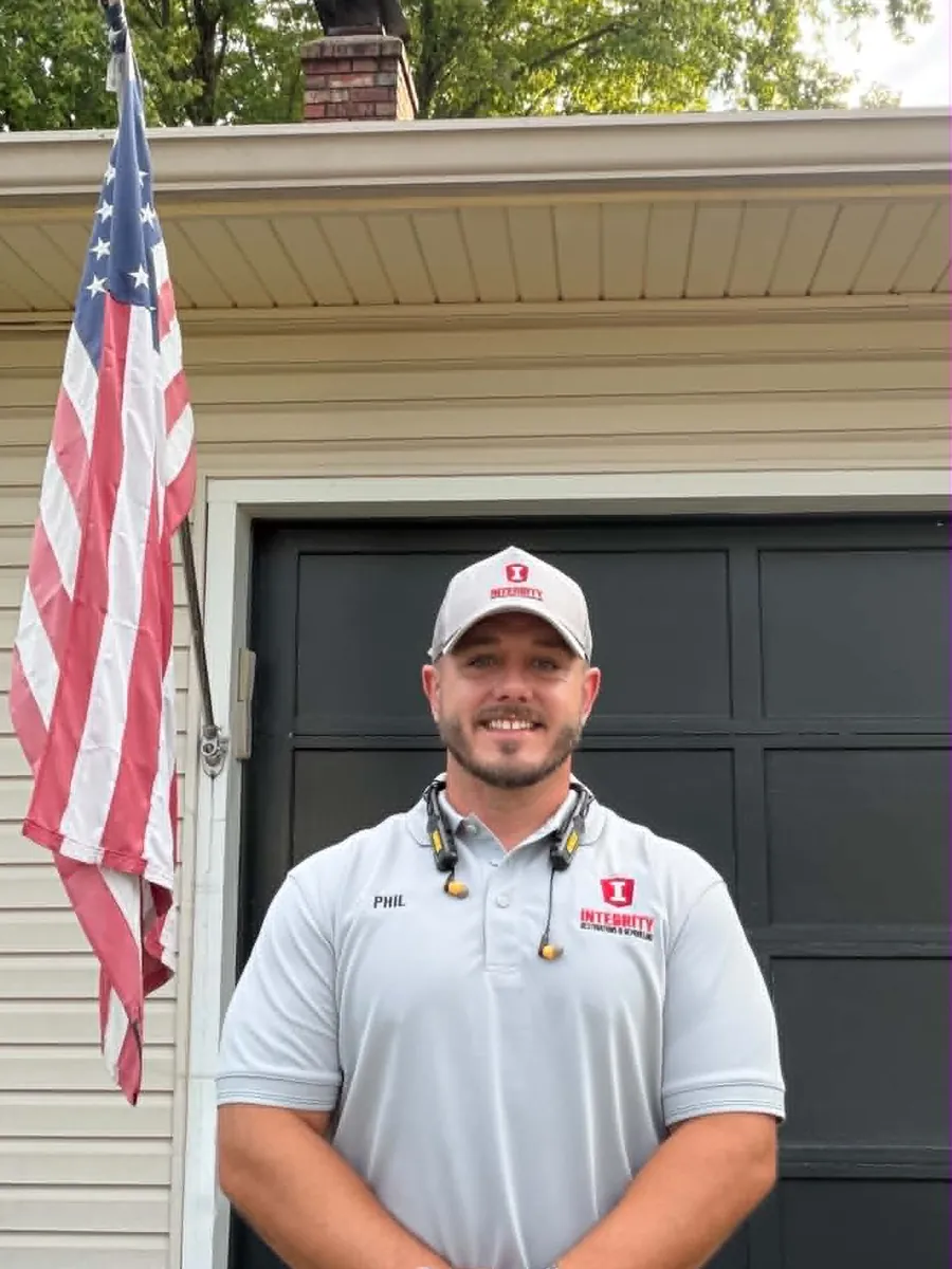Phil Betzner in an Integrity polo and cap, smiling in front of his garage with an American flag beside him