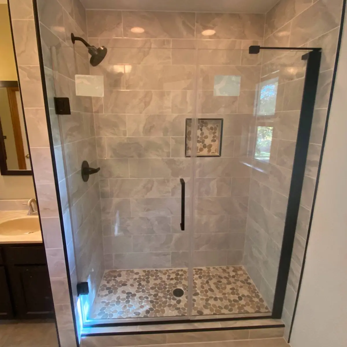 Same shower rebuilt: large-format tile, a niche, a pebble floor and a black-framed glass door
